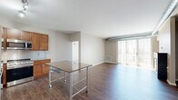 Located on Liberty Avenue in Pittsburgh's Bloomfield neighborhood, this three-bedroom, two-bathroom residence offers a well-designed single-floor layout with a thoughtful mix of living and functional spaces. The open-concept kitchen and living room serves as the heart of the home, where warm-toned wood cabinetry, granite countertops, stainless steel appliances — including a gas range, dishwasher, microwave, and refrigerator — and track lighting create an inviting and functional kitchen. Rich hardwood-style flooring extends throughout the main living area, which is anchored by a sleek, modern fireplace and opens directly to a private balcony, filling the space with natural light. The three bedrooms feature plush carpeting and neutral tones, with the generously sized primary bedroom offering ample room to furnish comfortably. Five closets throughout the unit provide exceptional storage. The two bathrooms are finished with granite-topped wood vanities and tile flooring; one includes a glass-enclosed shower while the other offers a full tub-shower combination with subway tile surround. A dedicated den adds flexibility for a home office or additional living space, and in-unit laundry adds everyday convenience. Exposed ductwork accents carry an urban, industrial character throughout the residence.