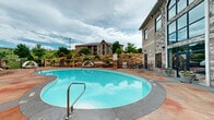 Located at 2710 Wilkie Dr in Rapid City, South Dakota, this outdoor amenity area is centered around a generously sized freeform swimming pool with crystal-clear blue water. The pool deck is finished in a rich, stamped concrete surface with warm earth tones, providing ample space for lounging and socializing. Surrounding the pool are cushioned lounge chairs, pod-style cabana seats, and bistro table-and-chair sets, offering a variety of relaxing and social settings.

A hot tub is conveniently positioned adjacent to the main pool, ideal for relaxation. The perimeter of the deck is enclosed by a decorative wrought iron fence accented with stone pillars, lending a polished, resort-like aesthetic. A built-in grill station and a vending machine add functional convenience to the outdoor experience.

The facility building features a striking stone and masonry exterior with large, arched windows that provide an architectural focal point. Lush landscaping with mature trees and natural rock formations frames the space beautifully. Beyond the fencing, sweeping views of the rolling green hills characteristic of the Black Hills region of South Dakota create a picturesque natural backdrop, enhancing the overall outdoor ambiance of this well-appointed recreational space.