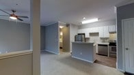 This single-floor residence showcases an open-concept design that seamlessly connects the living area and kitchen. The living space is finished with neutral-toned carpet, while the kitchen transitions to hard flooring, creating a clear yet fluid distinction between the two areas. The kitchen is well-appointed with white upper cabinetry, a peninsula-style counter that doubles as a casual dining or workspace, stainless steel appliances including a refrigerator and dishwasher, and a sink positioned along the perimeter counter. Recessed and flush-mounted ceiling lighting provides consistent illumination throughout the kitchen zone. The walls are painted in a cool, muted gray tone that complements the white cabinetry and neutral flooring. Crown molding adds a refined architectural detail along the upper wall edges. A hallway visible from the main living area leads to additional rooms, suggesting a functional layout designed for comfortable day-to-day living. The overall ambiance is clean, modern, and move-in ready, with a cohesive color palette that lends the space a calm, understated aesthetic.