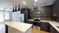 This single-floor space showcases a thoughtfully designed interior with a distinctly modern aesthetic. The kitchen serves as a central highlight, featuring rich, dark espresso-toned cabinetry that extends from floor to ceiling, maximizing storage capacity and creating a bold visual statement. Complementing the cabinetry are clean white quartz countertops that provide a striking contrast, along with a decorative mosaic tile backsplash that adds texture and visual interest behind the cooking area. Stainless steel appliances, including a French door refrigerator with an ice and water dispenser, a range with electric cooktop, and an over-the-range microwave, lend the kitchen a polished, professional quality. A spacious kitchen island with a white countertop offers additional workspace and casual seating potential. Three sleek matte black pendant lights hang above, providing focused task lighting while enhancing the contemporary design language. Light hardwood-style flooring runs throughout, warming the overall palette and grounding the space. A dedicated built-in wine rack adds a refined, functional touch. The open layout and carefully curated finishes throughout combine to create a cohesive, upscale living environment that is both practical and visually sophisticated.