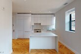 This thoughtfully designed single-floor residence combines modern aesthetics with functional living. The open-plan kitchen and living area serves as the heart of the home, showcasing striking herringbone-patterned hardwood floors that run throughout the entire space. The kitchen is fitted with floor-to-ceiling flat-front white cabinetry, a gas range, integrated appliances, and a clean white quartz island that doubles as a dining or prep surface. Recessed lighting illuminates the space, while black-framed windows allow natural light to flow in freely.

The living area is generously proportioned, offering an open and airy feel with soft neutral walls and continued herringbone flooring. Multiple bedrooms provide comfortable private retreats, each benefiting from large black-framed windows with views of the surrounding brick architecture, and the same warm herringbone flooring underfoot.

The residence includes two well-appointed bathrooms. One features a soaking tub with a glass enclosure and floor-to-ceiling tile work, while the second offers a frameless glass walk-in shower. Both bathrooms are finished with floating white vanities, large mirrors, and neutral stone-toned tile, creating a clean, spa-like atmosphere. A louvered interior door and a solid entry door with a keypad lock complete the practical, polished layout.