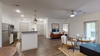 This single-floor residence showcases a thoughtfully designed open-concept layout that seamlessly connects the living, dining, and kitchen areas. Rich hardwood-style flooring flows throughout the entire space, anchoring the modern mid-century aesthetic. The expansive living room features vaulted ceilings, a ceiling fan, large windows with floor-length drapes that invite natural light, and direct access to an outdoor area via a glass door. The kitchen is outfitted with white shaker cabinetry, granite countertops, stainless steel appliances, and a peninsula with pendant lighting overhead — ideal for casual dining or entertaining. A dedicated dining nook accommodates a round wood table with seating for four, accompanied by a bold piece of wall art that adds character. The bedroom is generously proportioned, complete with a ceiling fan, a window, and ample wall space. The bathroom features white cabinetry with granite countertops, a single-basin sink, a large mirror, and a tub-shower combination. Throughout the residence, clean white walls, consistent flooring, and a cohesive neutral color palette with warm accent tones create a bright, welcoming, and well-organized living environment.