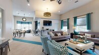 This well-appointed single-floor community space is designed to accommodate a variety of activities, offering distinct functional zones within an open, cohesive layout. A bold teal and neutral color scheme—expressed through patterned accent chairs, curtains, and carpet inlays—ties the entire space together and gives it a modern, polished character.

The central lounge area serves as the heart of the space, featuring comfortable seating arrangements including a gray sofa, teal-patterned armchairs, and a coffee table, all anchored beneath a decorative drum pendant light and ceiling fans. A wall-mounted flat-screen television above a textured accent wall creates a natural focal point for media viewing.

Adjacent to the lounge is a dedicated business center, equipped with desk workstations and task chairs, illuminated by recessed lighting and ample natural light from multiple windows. Tall interior windows with white trim connect this zone visually to a transitional corridor area, which features elevated ceilings and an arcade game console, adding a recreational touch.

A separate dining and lounge nook provides high-top bistro tables and bar seating alongside a bay window with teal valances, offering a casual gathering spot. Throughout the space, large windows dressed in teal curtains invite natural light and views of the surrounding exterior. The neutral carpet flooring with teal stripe accents and clean white walls contribute to an atmosphere that is both functional and inviting.