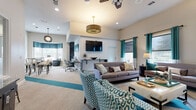 This well-appointed single-floor community space is designed to accommodate a variety of activities, offering distinct functional zones within an open, cohesive layout. A bold teal and neutral color scheme—expressed through patterned accent chairs, curtains, and carpet inlays—ties the entire space together and gives it a modern, polished character.

The central lounge area serves as the heart of the space, featuring comfortable seating arrangements including a gray sofa, teal-patterned armchairs, and a coffee table, all anchored beneath a decorative drum pendant light and ceiling fans. A wall-mounted flat-screen television above a textured accent wall creates a natural focal point for media viewing.

Adjacent to the lounge is a dedicated business center, equipped with desk workstations and task chairs, illuminated by recessed lighting and ample natural light from multiple windows. Tall interior windows with white trim connect this zone visually to a transitional corridor area, which features elevated ceilings and an arcade game console, adding a recreational touch.

A separate dining and lounge nook provides high-top bistro tables and bar seating alongside a bay window with teal valances, offering a casual gathering spot. Throughout the space, large windows dressed in teal curtains invite natural light and views of the surrounding exterior. The neutral carpet flooring with teal stripe accents and clean white walls contribute to an atmosphere that is both functional and inviting.