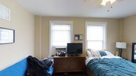 This single-floor bedroom presents a functional and lived-in layout with warm yellow-toned walls and crisp white trim framing two windows fitted with white horizontal blinds, allowing for adjustable natural light. The room is anchored by a substantial dark wood dresser positioned between the windows, which serves as both storage and an entertainment surface, currently holding a flat-screen television and various personal items. A window-mounted air conditioning unit provides climate control. The bed, positioned along the right wall near the second window, is dressed in blue bedding and adds a casual, comfortable feel to the space. Overhead lighting provides additional illumination, complementing the natural light that filters through the windows. The walls' soft yellow hue gives the room a warm, inviting ambiance, while the white ceiling maintains a sense of openness. The classic window trim and traditional dresser styling lend a slightly vintage character to the overall aesthetic. The room offers a modest but practical layout with enough space for essential furnishings and everyday living.