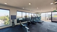 Located at 1250 N Fairfax Ave in West Hollywood, California, this well-appointed fitness center offers a comprehensive workout environment designed for serious training. The space is defined by its floor-to-ceiling windows along multiple walls, flooding the interior with abundant natural light and framing sweeping views of the surrounding West Hollywood skyline and iconic palm trees — creating an inspiring backdrop for every workout.

The facility is equipped with a full lineup of True-brand cardio machines, including treadmills, ellipticals, and a stationary air bike, all positioned to take advantage of the outdoor views. A dedicated rowing machine rounds out the cardio offerings. For strength training, the space features a large functional training cable rig, a comprehensive free weights area with a full dumbbell rack, and an assortment of benches and accessories. A heavy bag is also available for boxing and conditioning work.

Underfoot, commercial-grade rubber flooring in a dark tile pattern provides durability and impact absorption throughout the space. Full-length mirrors are mounted along interior walls, enhancing the sense of openness and aiding in form monitoring during training. Recessed lighting supplements the natural light for consistent illumination. A sliding glass door connects the fitness floor to an adjacent outdoor terrace, offering direct access to fresh air. The overall design is clean, modern, and purposefully outfitted for a high-quality fitness experience.