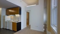 This single-floor space showcases a clean, modern design with a thoughtfully open layout that connects the kitchen and living areas seamlessly. The kitchen is outfitted with sleek dark cabinetry contrasted by warm wood-toned upper cabinets, a stainless steel sink with a contemporary gooseneck faucet, and under-cabinet lighting that adds a functional yet stylish touch. A linear ceiling-mounted light fixture illuminates the kitchen area, contributing to the bright, airy atmosphere throughout. The walls are finished in crisp white, creating a neutral and versatile backdrop that enhances the sense of openness. The living area features soft wall-to-wall carpeting, offering comfort underfoot. An open doorway leads to an additional room or hallway, suggesting a well-connected floor plan that promotes easy movement through the space. The elevated ceiling height adds a sense of volume and spaciousness, making the interior feel larger than its footprint. Overall, the space presents a minimalist aesthetic with practical design choices, ideal for those who appreciate a clean, uncluttered environment with modern finishes.