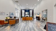 This well-appointed single-floor community space offers a thoughtfully designed open-concept layout that combines welcoming common areas with functional amenities. Upon entry, soaring ceilings and floor-to-ceiling glass doors immediately create a bright, airy ambiance, with abundant natural light streaming throughout the space. The flooring throughout features wide-plank wood-look planks in cool grey tones, lending a contemporary and cohesive aesthetic.

The main lounge area serves as the centerpiece, anchored by a classic white fireplace set against a striking navy blue accent wall flanked by tall windows with dark drape treatments. Comfortable seating — including a rich cognac leather sectional and tufted blue accent chairs — creates an inviting atmosphere for gathering. A dedicated reception or leasing desk area with warm wood tones occupies one side, while a bar-height table and cabinet provide an additional social zone.

A glass-partitioned fitness room is visible from the main seating area, housing cardio equipment and exercise bikes, and adding a sense of transparency and openness to the overall design. Crown molding, recessed lighting, and carefully placed artwork and greenery contribute to the polished, modern-yet-welcoming character of this versatile community space.
