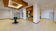 This single-floor space in Oakland, CA presents a thoughtfully designed communal environment that blends contemporary architecture with warm, natural materials. The open layout is anchored by light-toned large-format tile flooring that extends throughout, creating a cohesive and airy atmosphere.

The central reception or lobby area draws immediate attention with a striking coffered ceiling element clad in rich warm wood, from which a sculptural multi-pendant brass and gold chandelier descends, casting an elegant ambient glow. A bold pedestal table with decorative greenery serves as a focal centerpiece beneath this feature ceiling.

Built-in warm wood shelving units are integrated into the structural columns, providing both display space and architectural interest. Curated sculptural objects and decorative pieces are arranged throughout these shelves, lending a gallery-like quality to the space.

An adjacent lounge area features a dramatic backlit wood-paneled accent wall with a geometric chevron pattern, complemented by a long upholstered built-in bench and plush swivel armchairs in cognac velvet, creating an inviting seating nook. A separate, naturally lit room accessible through accordion-style doors offers additional lounge seating with floor-to-ceiling windows. Black-and-white framed photography adorns the corridor walls, reinforcing the space's curated, design-forward character.