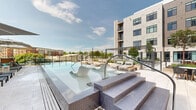 Located at 115 Coulter Ave in Ardmore, Pennsylvania, this outdoor amenity deck is set within a modern multi-story residential building and offers a thoughtfully designed communal retreat. The centerpiece of the space is a generously sized resort-style swimming pool, complete with built-in in-water lounge chairs that invite relaxation directly in the water. The pool deck is finished in large-format neutral pavers, providing a clean and contemporary aesthetic that complements the building's modern facade of light-toned panels and expansive glass windows.

Surrounding the pool, multiple seating zones are arranged to accommodate a variety of outdoor activities. Cushioned chaise lounges and modular sectional sofas with teak accents and sage green accent pillows create comfortable gathering areas, while large market umbrellas offer ample shade throughout the day. A covered pergola anchors a dedicated lounge and entertainment zone, featuring string lights overhead and views of the pool and landscaping beyond.

Lush plantings — including ornamental grasses, lavender, and young trees — are interwoven throughout the deck, softening the hardscape and adding natural color and texture. Glass-panel railings along the perimeter preserve unobstructed sightlines to the surrounding Ardmore neighborhood and open sky. The overall ambiance is one of polished urban resort living, blending refined outdoor design with a welcoming, communal atmosphere.
