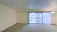 This single-floor space presents a straightforward, open layout centered around a generously sized living area. The room is finished with wall-to-wall carpet in a neutral tone, providing a soft underfoot feel throughout the space. A prominent feature is the expansive floor-to-ceiling sliding glass door fitted with vertical blinds, which spans a significant portion of one wall and allows abundant natural light to fill the interior. The door opens to a balcony that overlooks an exterior courtyard or neighboring building, offering a connection to the outdoors. The walls are painted in a light, neutral hue, contributing to an airy and bright atmosphere. The ceiling features a subtle textured finish, adding a modest layer of visual detail to the overhead space. The overall design is clean and minimal, with smooth, unadorned walls that provide flexibility for personalization. The open nature of the room allows for a variety of furniture arrangements, making the space adaptable to different functional needs. The combination of natural light, neutral finishes, and open layout creates a calm and practical living environment.