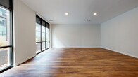 This contemporary two-floor residence showcases a thoughtfully designed layout that balances open, airy living spaces with functional, well-finished interiors. The main living area is a standout, featuring rich, wide-plank hardwood flooring and floor-to-ceiling windows with black-framed sliding panels that invite an abundance of natural light and offer views of the surrounding urban streetscape. Recessed lighting complements the clean white walls and smooth ceilings throughout.

The kitchen is a highlight of the space, offering dark wood-grain cabinetry with sleek bar-pull hardware, a white subway tile backsplash, and generous stone countertops with subtle veining. A large peninsula with a waterfall stone edge and an integrated sink provides both additional workspace and a casual gathering point. Stainless steel appliances, including a refrigerator, electric range, microwave, and dishwasher, complete this modern culinary space.

The bathrooms are finished with large-format light tile flooring and warm wood-toned vanities topped with stone counters. One bathroom features a full tub-shower combination with tiled surround, while a second offers a more compact layout with overhead storage cabinetry. Carpeted bedrooms provide a softer contrast to the hardwood common areas, with windows drawing in natural light. A utility or storage room with locker-style cabinetry and carpet adds practical versatility to the overall floor plan.