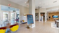 This single-floor community clubhouse at Dream Kissimmee offers a thoughtfully designed and generously proportioned space that blends modern aesthetics with a welcoming atmosphere. The main entry lobby immediately impresses with soaring ceilings, structural columns, and light wood-toned flooring that extends throughout the facility. Recessed lighting and ceiling fans contribute to the bright, airy ambiance, while large glass entry doors invite natural light into the open corridor.

The lounge area is furnished with contemporary blue and neutral-toned seating, accent tables, and a large wall-mounted television, complemented by decorative metalwork wall art and curated greenery. A separate, more intimate sitting room features a curved sectional sofa, abstract artwork, and a wall-mounted display screen, creating a cozy yet stylish retreat.

A dedicated coffee bar area provides residents with white shaker cabinetry, granite countertops, a stainless steel refrigerator, a blue tile backsplash, and a Starbucks-branded coffee machine. A distinctive foyer vignette near the interior corridor showcases a sculptural glass-top accent table with a dramatic dried pampas grass arrangement, adding an artistic focal point to the transitional space.