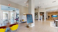 This single-floor community clubhouse at Dream Kissimmee offers a thoughtfully designed and generously proportioned space that blends modern aesthetics with a welcoming atmosphere. The main entry lobby immediately impresses with soaring ceilings, structural columns, and light wood-toned flooring that extends throughout the facility. Recessed lighting and ceiling fans contribute to the bright, airy ambiance, while large glass entry doors invite natural light into the open corridor.

The lounge area is furnished with contemporary blue and neutral-toned seating, accent tables, and a large wall-mounted television, complemented by decorative metalwork wall art and curated greenery. A separate, more intimate sitting room features a curved sectional sofa, abstract artwork, and a wall-mounted display screen, creating a cozy yet stylish retreat.

A dedicated coffee bar area provides residents with white shaker cabinetry, granite countertops, a stainless steel refrigerator, a blue tile backsplash, and a Starbucks-branded coffee machine. A distinctive foyer vignette near the interior corridor showcases a sculptural glass-top accent table with a dramatic dried pampas grass arrangement, adding an artistic focal point to the transitional space.