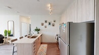 Located at 914 S Milton Rd in Flagstaff, Arizona, this thoughtfully designed unit offers four bedrooms and four bathrooms across a single floor, making it well-suited for shared living arrangements. The open-concept kitchen and living area serves as the heart of the home, featuring a spacious island with bar seating, light wood cabinetry, stainless steel appliances including a refrigerator, electric range, and microwave, all illuminated by track lighting overhead. The adjoining living room is bright and inviting, benefiting from a large window dressed with sheer white curtains that allow abundant natural light to fill the space. Wood-look plank flooring flows consistently throughout, adding warmth and visual continuity. Each bedroom is privately scaled, offering window-facing natural light, closet storage — some with mirrored sliding doors — and enough room for a bed and study desk, making them functional for student or professional living. Every bedroom is paired with its own en-suite bathroom, each finished with a modern floating vanity, light wood cabinetry, white countertops, and large mirrors. The clean, neutral color palette of white and warm beige tones is carried throughout the entire unit, creating a cohesive and contemporary aesthetic. A hallway connects the private bedrooms to the shared living spaces, ensuring a practical and well-organized layout.