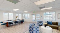 Located at 21620 Dracaea Avenue in Moreno Valley, CA, this single-floor community clubhouse offers a well-appointed gathering space designed for both relaxation and socializing. The interior is finished with light wood-look flooring, crisp white walls, and a drop ceiling with integrated panel lighting that keeps the space bright and welcoming throughout.

The dining area features a large wood dining table surrounded by plush blue upholstered chairs, bathed in natural light from multiple windows fitted with white horizontal blinds. Coastal-inspired botanical wall art and a statement dried-grass arrangement add tasteful decorative touches.

Adjacent to the dining area, a comfortable lounge zone is furnished with a sectional sofa in warm olive tones, accented by blue patterned armchairs and a round white coffee table — creating an inviting setting for casual conversation. Glass double doors open directly to an outdoor pool area, allowing natural light to pour in and offering easy access to exterior amenities.

The game room is a standout feature, centered around a full-size pool table with a navy felt surface. A geometric-patterned accent wall serves as a striking backdrop for a wall-mounted flat-screen TV, while bar-height seating and framed artwork complete the space. The overall ambiance is clean, modern, and community-focused.