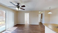 This single-story residence is thoughtfully laid out to maximize functionality across its living spaces. The open living area serves as the central hub of the home, showcasing rich wood-style flooring that extends throughout the main areas. A ceiling fan with integrated light fixture is mounted to the textured ceiling, providing both comfort and illumination. A sliding glass door allows natural light to pour into the living space while providing direct access to an outdoor patio area. A pass-through countertop with granite-style surfaces connects the living area to the kitchen, creating an open, communicative flow between spaces.

The galley-style kitchen is efficiently designed with warm-toned wood cabinetry lining both walls, offering generous storage. Granite-style countertops provide ample prep space, and white appliances including a refrigerator, dishwasher, and range are included. The wood-style flooring continues seamlessly into the kitchen.

The bedroom is carpeted for comfort and features a ceiling fan, a wide window fitted with blinds that allows natural light while maintaining privacy, and neutral wall tones throughout. The textured ceilings are consistent across all rooms, contributing to a cohesive aesthetic throughout the residence.