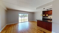 Located at 21 Kristin Dr in Schaumburg, Illinois, this single-floor residence offers a thoughtfully designed open-concept layout that seamlessly connects the living and kitchen areas. Warm-toned hardwood flooring runs throughout the main living spaces, providing a cohesive and inviting foundation. The living area is generously sized and bathed in natural light, courtesy of large sliding glass doors that open to outdoor views and bring a bright, airy quality to the interior. Vaulted ceilings in portions of the main living space add a sense of vertical openness. The kitchen is a standout feature, outfitted with rich cherry wood cabinetry, dark granite countertops, stainless steel appliances — including a refrigerator and dishwasher — and a central island with pendant lighting overhead. Track lighting above the kitchen provides additional illumination. A secondary room with its own sliding glass door offers flexible use as a bedroom or office, with the same hardwood flooring continuing throughout. The bathroom is finished with full-height warm-toned tile surrounding a tub-shower combination, complemented by tile flooring. Neutral gray walls throughout the space create a clean, modern backdrop. The textured ceiling adds a classic touch, while the overall layout prioritizes functionality and comfort.