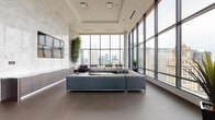 Located in the heart of Chicago's Loop at 73 E Lake Street, this upscale lounge space offers a refined, contemporary environment elevated high above the city. The open-plan layout is anchored by dramatic floor-to-ceiling curtain-wall windows that frame sweeping panoramic views of Chicago's iconic skyline, including prominent landmarks such as Trump Tower. Natural light floods the interior throughout the day, creating a bright and inviting atmosphere.

The design language is clean and modern, with large-format porcelain tile flooring in warm taupe tones providing a neutral foundation. A striking full-height marble-clad feature wall commands attention, lending the space a sense of luxury and permanence. Recessed ceiling lighting and a tray ceiling detail add architectural depth overhead.

The lounge is furnished with generously sized sectional sofas and plush velvet armchairs in sophisticated grey tones, arranged to encourage both social interaction and individual relaxation. A built-in credenza with wood-grain cabinetry runs along one wall, accompanied by a wall-mounted multi-panel display and integrated countertop with sink, providing practical amenities within an elegant setting. A high-top bar counter with sleek stools offers additional gathering space. Tall indoor trees introduce organic warmth and contrast beautifully against the polished marble and glass surfaces throughout.