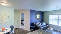 This single-floor residence offers a thoughtfully designed living space that balances style and comfort. The living area immediately draws attention with its striking navy blue accent wall, adorned with a decorative sunburst mirror that serves as a bold focal point. A dark leather sofa, accented with an assortment of patterned and solid throw pillows in coordinating blue and neutral tones, anchors the seating area. A small round side table adds a functional yet decorative touch. The flooring throughout the main living space consists of rich, dark hardwood-style planks, lending warmth and a modern aesthetic to the room. The walls are finished in soft, neutral tones that complement the vibrant accent wall, while smooth white ceilings contribute to a clean, bright atmosphere. An open doorway connects the living area to a carpeted bedroom, where a neatly made bed and soft, muted décor create a calm and restful retreat. The light-toned carpet in the bedroom provides a comfortable contrast to the harder flooring in the main living space. Overall, the home presents a cohesive interior design with a pleasing balance of bold color choices and neutral backdrops, creating an inviting and well-appointed environment.