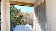This single-floor residence offers a comfortable and functional layout with several notable features throughout. The main living area is carpeted and anchored by a tile-surround fireplace, creating a warm and inviting focal point. A ceiling fan with integrated lighting overhead adds both comfort and practicality, while a textured ceiling lends a classic residential feel. A pass-through window connects the living area to the kitchen, which is equipped with white appliances including a refrigerator and range, and features cabinetry with a dark-toned valance above. This open connection between the kitchen and living space promotes an easy flow and makes the area feel more expansive. A hallway visible from the main room leads further into the residence, suggesting additional private spaces beyond. The space also includes a private outdoor balcony accessible via a sliding door secured with a decorative wrought-iron security door. The balcony features metal railings and offers pleasant views of mature trees, lush landscaping, and the surrounding stucco-exterior buildings under open skies. This outdoor space provides a peaceful retreat and extends the overall livable area of the residence.