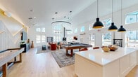 Located at 14378 Walnut Grove Way in Oregon City, Oregon, this single-floor community living space offers a generously sized, open-plan layout designed for social interaction and relaxation. The room's most striking architectural feature is its soaring vaulted ceiling, which is lined with a band of clerestory windows that flood the interior with abundant natural light throughout the day. Wide-plank light hardwood flooring runs throughout, contributing to a warm, cohesive aesthetic.

The space is organized into several distinct functional zones. A central lounge area features plush, vibrantly colored seating—including bold red accent chairs—arranged around layered area rugs, with a freestanding fireplace adding warmth and character. A full-size pool table anchors the middle of the room beneath a dramatic circular pendant light fixture. Adjacent to this is a kitchenette area outfitted with a striking navy blue tile backsplash, bar-height seating, and pendant lighting.

A dedicated media area showcases a large wall-mounted television set against a distinctive wood-slat accent wall with an angled yellow ceiling detail and integrated LED backlighting. A built-in window bench with outdoor views and a wall-mounted oversized Scrabble board add playful, community-focused touches. The overall ambiance is modern, bright, and inviting.