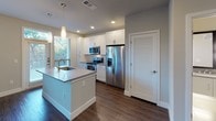 This well-designed single-floor residence showcases a clean, modern aesthetic with an open-concept layout that seamlessly connects the living, dining, and kitchen areas. Rich, dark hardwood-style flooring runs throughout the main living spaces, lending warmth and continuity to the interior.

The kitchen is a standout feature, equipped with crisp white shaker-style cabinetry extending to the ceiling for ample storage, stainless steel appliances including an electric range, over-the-range microwave, and a French door refrigerator. A decorative tile backsplash adds texture, while a central island with a light quartz countertop and undermount sink provides both workspace and casual seating. Pendant lighting above the island and recessed ceiling lights complete the polished look.

The living area is bright and airy, benefiting from a large bank of windows that invite abundant natural light and views of surrounding greenery. A ceiling fan with an integrated light fixture adds comfort and functionality. An adjacent room, accessible through an open pass-through, offers flexible use as a bedroom or secondary living space, also finished with matching flooring and a ceiling fan.

The bathroom is finished in a neutral palette with large-format floor tiles, a quartz-topped vanity, wall-mounted sconce lighting, and a full tub-shower combination with a tiled surround and built-in niche.