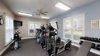 This single-level fitness center offers a comprehensive workout environment designed to accommodate a variety of exercise needs. The space is laid out in an elongated rectangular floor plan, providing ample room to house an extensive selection of equipment without feeling cramped.

The cardio zone is prominently featured and includes multiple elliptical trainers, treadmills, and a recumbent bike, all arranged to take advantage of the generous natural light pouring in through numerous white-trimmed windows fitted with horizontal blinds. Views of the surrounding outdoor amenities, including a pool area and landscaped grounds, are visible through the glass-paneled double entry doors and surrounding windows.

A dedicated strength and free-weight area features a tiered dumbbell rack, a flat workout bench, a cable machine, and a wall-mounted battle rope station. A wall-mounted flat-screen television is positioned near the cardio equipment for entertainment during workouts.

The interior finishes include durable rubber flooring throughout, light blue and warm beige painted walls, and flush-mounted fluorescent lighting panels. Ceiling fans help maintain comfortable air circulation. Safety features such as a fire extinguisher, hand sanitizer dispenser, and exit signage are also present, reflecting a thoughtfully maintained and functional fitness facility.