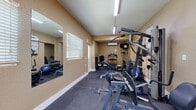 Located in Seminole, Florida, this dedicated fitness center offers a well-equipped exercise space designed to support a comprehensive workout experience. The room features vaulted ceilings that lend a sense of openness, with warm beige walls and durable rubber mat flooring throughout, providing both safety and comfort during exercise.

The space is outfitted with a multi-station cable and weight machine, a leg press, an adjustable workout bench, and a dumbbell rack with a full range of free weights. A stability ball adds flexibility training options. On the cardio side, a treadmill and an elliptical trainer are positioned at the far end of the room, accompanied by a wall-mounted flat-screen television for entertainment during workouts.

Climate control is handled by a wall-mounted mini-split air conditioning unit, ensuring comfort year-round. Natural light enters through a window with blinds, complemented by overhead fluorescent lighting. A large wall mirror along one side enhances the sense of space and allows users to monitor their form. A water cooler is available for hydration convenience.

Safety and informational signage, including fitness center rules and exercise reference charts, are posted on the walls. The space is accessed through a secured entry door and is equipped with an illuminated exit sign, reflecting proper safety standards.
