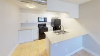 This single-floor unit offers a practical and comfortable layout designed to maximize functionality throughout. The kitchen is a standout feature, equipped with white shaker-style cabinetry, light countertops with a tile backsplash, and a full suite of black appliances including a gas range, built-in microwave, and refrigerator. A peninsula with a stainless steel sink provides additional prep space and a natural transition into the adjoining living area. The living space is finished with light ceramic tile flooring and neutral gray walls, creating a bright and open atmosphere. A window fitted with horizontal blinds allows natural light to filter in while offering privacy and a view of surrounding greenery. The bathroom is thoughtfully updated with a white vanity, framed mirror, light countertop, and wood-look vinyl plank flooring that adds warmth to the clean, crisp aesthetic. Grab bars and a towel bar provide practical convenience. A dedicated laundry area with a stacked washer and dryer unit offers the convenience of in-unit washing, situated adjacent to the bathroom with additional shelving for storage. The overall design palette of whites, grays, and warm wood tones creates a cohesive and inviting living environment throughout the space.
