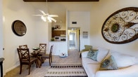 This single-floor residence offers a welcoming and functional layout designed for comfortable everyday living. The open-concept main area seamlessly blends the living room, dining space, and kitchen into one cohesive environment. The living room is outfitted with plush carpeting and benefits from vaulted ceilings accented by exposed dark wood beams, lending a warm architectural character to the space. A ceiling fan provides both comfort and a casual charm. Natural light filters through the space, enhancing its bright and airy atmosphere. The dining area sits conveniently adjacent to the living room, providing an intimate setting for meals. The kitchen features dark wood cabinetry and is partially open to the main living area via a pass-through counter, making it easy to interact with guests while preparing food. A hallway visible from the main living area leads to additional rooms, suggesting a practical separation between the public and private areas of the home. Soft, neutral tones throughout the walls and flooring create a calm and inviting ambiance, while decorative accents add a personalized touch. Overall, this residence balances functional design with a cozy, homey aesthetic suited for comfortable day-to-day living.