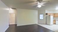 This single-floor space offers a clean, straightforward layout designed for comfortable everyday living. The main living area is defined by rich, dark hardwood-style flooring that contrasts elegantly with the bright, neutral-toned walls, creating a warm yet modern aesthetic. A white five-blade ceiling fan with an integrated light fixture is centrally mounted, providing both air circulation and ambient lighting to the space. The front entry door features a multi-pane window that allows natural light to filter in, brightening the interior and offering a welcoming transition from the outside. A standard doorway on the opposite side of the room leads to additional areas of the home, suggesting a functional flow between spaces. The walls are finished in a soft, off-white tone that keeps the space feeling open and airy. A ceiling-mounted HVAC vent ensures year-round climate control. The overall ambiance is modest and neutral, providing a versatile canvas well-suited for a variety of interior styles and furnishing arrangements. The layout prioritizes simplicity and practicality, making it an accessible and easy-to-navigate single-level space.