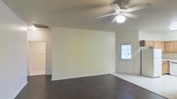 This single-floor space offers a clean, straightforward layout designed for comfortable everyday living. The main living area is defined by rich, dark hardwood-style flooring that contrasts elegantly with the bright, neutral-toned walls, creating a warm yet modern aesthetic. A white five-blade ceiling fan with an integrated light fixture is centrally mounted, providing both air circulation and ambient lighting to the space. The front entry door features a multi-pane window that allows natural light to filter in, brightening the interior and offering a welcoming transition from the outside. A standard doorway on the opposite side of the room leads to additional areas of the home, suggesting a functional flow between spaces. The walls are finished in a soft, off-white tone that keeps the space feeling open and airy. A ceiling-mounted HVAC vent ensures year-round climate control. The overall ambiance is modest and neutral, providing a versatile canvas well-suited for a variety of interior styles and furnishing arrangements. The layout prioritizes simplicity and practicality, making it an accessible and easy-to-navigate single-level space.