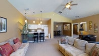 This single-floor residence showcases an open-concept layout that seamlessly connects the living, dining, and kitchen areas into one cohesive, flowing space. The most striking architectural feature is the vaulted ceiling, which dramatically increases the sense of vertical space and openness throughout the main living area. A ceiling fan is centrally mounted, providing both function and a finishing touch to the soaring roofline.

The kitchen is well-appointed with white cabinetry, white appliances including a full-size refrigerator, and a dark-topped breakfast bar with seating for multiple guests. Pendant lighting hangs above both the kitchen and dining areas, adding warmth and definition to each zone. The dining area accommodates a full table and chairs, making it ideal for entertaining.

The living room features neutral carpeting and is furnished with comfortable seating, accented by decorative throw pillows. Warm yellow-toned walls throughout give the space an inviting and cheerful ambiance. A large potted palm adds a natural, tropical element to the room. Additional rooms are visible beyond the main living area, connected by open doorways that maintain the home's airy, accessible feel.