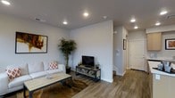 This well-appointed two-story space showcases a thoughtfully designed interior with a clean, modern aesthetic. The main living area features warm wood-look hardwood flooring that runs throughout, complementing the crisp white walls and smooth grey ceilings. Recessed lighting is strategically placed to provide even, ambient illumination across the space. The living room is open and welcoming, with ample room for comfortable seating and entertainment, centered around a sleek media console. Decorative touches, including framed wall art and potted indoor plants, add warmth and life to the neutral palette. A staircase leads to the second floor, hinting at additional living or sleeping quarters above. The layout flows naturally from the living area toward a hallway, which connects to additional rooms beyond. White interior doors and clean baseboards throughout maintain a polished, cohesive look. The overall ambiance is modern and understated, with a neutral color scheme that creates a versatile backdrop suitable for a variety of uses. The combination of natural-toned flooring, contemporary fixtures, and thoughtful spatial planning makes this a functional and visually appealing two-level space.