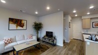 This well-appointed two-story space showcases a thoughtfully designed interior with a clean, modern aesthetic. The main living area features warm wood-look hardwood flooring that runs throughout, complementing the crisp white walls and smooth grey ceilings. Recessed lighting is strategically placed to provide even, ambient illumination across the space. The living room is open and welcoming, with ample room for comfortable seating and entertainment, centered around a sleek media console. Decorative touches, including framed wall art and potted indoor plants, add warmth and life to the neutral palette. A staircase leads to the second floor, hinting at additional living or sleeping quarters above. The layout flows naturally from the living area toward a hallway, which connects to additional rooms beyond. White interior doors and clean baseboards throughout maintain a polished, cohesive look. The overall ambiance is modern and understated, with a neutral color scheme that creates a versatile backdrop suitable for a variety of uses. The combination of natural-toned flooring, contemporary fixtures, and thoughtful spatial planning makes this a functional and visually appealing two-level space.