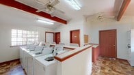 This single-story laundry facility is designed to efficiently serve a shared-use community. The main wash area houses a generous row of top-loading washing machines arranged along a central dividing counter with a red-trimmed surround, providing an organized and accessible layout. A dedicated dryer room along one side contains multiple stacked dryer units, maximizing capacity within a compact footprint. A separate alcove features an additional stacked washer-dryer unit alongside a broad folding counter positioned beneath a window, offering natural light and a practical workspace for sorting and folding laundry. Throughout the facility, the white walls are accented by bold red-painted exposed beams and door trim, lending a consistent visual identity to the space. Ceiling fans are mounted overhead to maintain comfortable airflow, complemented by fluorescent lighting that ensures the space is well-lit at all hours. The flooring throughout consists of durable stone-patterned tile, well-suited to withstand heavy foot traffic. Multiple entry doors provide convenient access from different points, and a corkboard is available for posting notices. Overall, the facility is functional, straightforward, and equipped to handle the laundry needs of a community.
