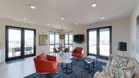 This single-floor space offers a seamlessly integrated living and dining area filled with natural light streaming through multiple large, dark-framed windows and glass doors that frame sweeping views of the surrounding skyline and rooftops. The open layout creates a generous, airy feel, enhanced by clean white ceilings outfitted with recessed lighting throughout.

The living area is anchored by striking red leather armchairs arranged around a navy patterned area rug, complemented by sleek black metal accent tables. A wall-mounted flat-screen television serves as a functional focal point, while the bold furnishings add a vibrant contrast to the neutral walls and light hardwood flooring.

Adjacent to the living space, the dining area features a round glass-top table surrounded by mid-century modern chairs in warm wood tones. A woven pendant light fixture hangs above, adding texture and warmth to the space.

Large glass doors along multiple walls provide direct access to an outdoor area, reinforcing the bright, connected ambiance. The overall design balances contemporary style with comfort, creating an inviting and well-lit environment suitable for both relaxing and entertaining.
