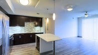This single-floor space showcases a thoughtfully designed open-concept layout that seamlessly connects the kitchen and living areas. The kitchen is outfitted with dark espresso cabinetry contrasted by a light-toned countertop island, which doubles as a casual dining and prep surface. A mosaic tile backsplash in neutral tones of beige, grey, and white adds visual texture behind the sink and counter workspace. Sleek stainless steel fixtures and integrated appliances, including a wine cooler beneath the island, contribute to the kitchen's modern aesthetic. Elongated pendant lights with a brushed nickel finish hang above the island, providing focused task lighting while enhancing the contemporary design. The flooring transitions between darker hardwood in the kitchen zone and lighter wood-toned planks in the adjacent living area, subtly defining each space. Smooth white walls and standard ceilings with recessed tray detailing create a clean, airy atmosphere throughout. The open flow between areas makes the space feel expansive and well-connected, ideal for both everyday living and entertaining.