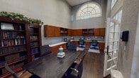 This single-floor business center is designed to support productivity and collaboration in an elegant, professional setting. The space is defined by its dramatically high ceilings, which create an open and airy atmosphere throughout. A striking arched transom window near the ceiling draws in generous natural light, illuminating the room's warm gray walls and rich dark hardwood flooring.

The room is divided into two functional zones. A central conference area features a large, dark wood table surrounded by ergonomic task chairs, making it well-suited for meetings or group work. French doors with glass transoms connect this area to an adjacent space, maintaining an open sightline while allowing for separation when needed.

Along the perimeter, a built-in L-shaped workstation constructed of warm honey-toned wood cabinetry provides dedicated computer workspaces equipped with monitors and a printer. Upper cabinets and open shelving offer ample storage and display space for books and reference materials. Freestanding dark wood bookshelves filled with volumes of books line one wall, topped with decorative greenery that adds a touch of warmth to the professional environment.

A cork bulletin board is mounted near the French doors for informational postings, and the overall layout reflects a thoughtfully organized space suited for both individual work and collaborative gatherings.