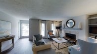 This single-floor residence offers a thoughtfully designed living space that balances comfort and style. The living room serves as the heart of the home, anchored by a tile-surround fireplace and framed by built-in shelving that provides both function and character. Large windows dressed with flowing sheer curtains flood the room with natural light, creating a bright and welcoming atmosphere throughout the day. The textured ceiling adds a layer of architectural detail, while the warm hardwood flooring grounds the space with a rich, natural tone. The interior is styled with a curated mix of contemporary furnishings, including a tufted sofa, an upholstered wingback chair, and a sleek marble-top coffee table set atop a cowhide area rug — all contributing to a sophisticated yet relaxed aesthetic. An octagonal wood-framed mirror adorns the fireplace wall, adding visual depth and a decorative focal point. The neutral gray wall palette provides a versatile backdrop that complements the warm accent tones found throughout the furnishings. Overall, this residence exudes a modern, inviting ambiance with attention to detail evident in every corner of the living space.