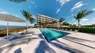 This outdoor amenity space presents a polished, resort-inspired atmosphere defined by its expansive rectangular pool with vivid turquoise water and a pristine light-toned tile surround. The pool deck is generously appointed with rows of sleek, modern sun loungers and shaded umbrella stations positioned along both sides, offering ample space for relaxation and leisure. Tall, mature palm trees frame the area, lending a distinctly tropical character that is enhanced by open views of the waterway visible along the property's edge. The backdrop features a well-maintained, contemporary multi-story building with clean horizontal lines, large balconied windows, and a crisp white facade that reflects the bright South Florida sunlight. The overall ambiance is bright, airy, and sophisticated, with the combination of lush landscaping, open sky, and shimmering water creating a serene and inviting outdoor environment. The meticulously maintained hardscape and carefully curated furnishings reinforce the upscale, resort-quality character of the space, making it an ideal setting for both quiet retreat and social gatherings in a sun-drenched waterfront setting.