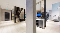 This two-floor fitness facility is designed with both function and aesthetics in mind, blending industrial and contemporary elements throughout. The entrance level greets visitors with light wood flooring, exposed concrete columns, and a dramatic built-in aquarium set within a wood-paneled wall, establishing an upscale tone from the outset. A sculptural open staircase with dark metal railings and vertical wood balusters connects the two levels, serving as a visual centerpiece.

The main gym floor is expansive and open, featuring exposed wood beam ceiling details, whitewashed brick accent walls, and the bold phrase "Feel the Burn" displayed prominently. The space is outfitted with a comprehensive selection of strength and cardio equipment, including stair climbers, cable machines, free weights, and functional training rigs. Large mirrors and windows allow natural light to fill the space. A turf-lined functional training corridor and a dedicated studio room with mirrored walls, light wood flooring, and a floor projector offer versatile workout options. The second level continues the gym programming, with glass-partitioned training zones and open views to the floor below. A marble-tiled shower room with glass enclosure provides post-workout amenities.