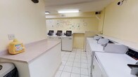 This communal laundry facility is a practical, well-organized space designed to accommodate multiple users at once. The room is finished with light yellow walls and clean white tile flooring, creating a bright and functional environment. Overhead fluorescent lighting ensures the space is well-lit throughout.

A charming "The Laundry Room" wall graphic with a bubble motif serves as a decorative focal point on the back wall, adding a touch of personality to the otherwise utilitarian space. Multiple top-loading washing machines are neatly arranged along the back wall, each equipped with digital control panels for ease of use. Additional machines — including what appear to be dryers — line the right side of the room, providing ample capacity for simultaneous loads.

A long, smooth folding counter runs along the left side of the room, offering generous surface space for sorting and folding laundry. Ventilation is handled via wall-mounted HVAC vents, keeping the room comfortable during use. The layout is thoughtfully arranged to allow easy movement between machines and the folding area, making the overall laundry experience efficient and straightforward.