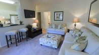 Located at 5454 Crossroads Crest Way in Cary, North Carolina, this single-floor residence offers a thoughtfully designed living environment with a welcoming and comfortable atmosphere. The living room serves as the heart of the home, featuring soft carpeted flooring and notably elevated ceilings that contribute to an airy, open feel. Warm, neutral wall tones create a calming backdrop throughout the space. The room is well-suited for both relaxation and entertaining, with ample space for seating arrangements and media. A wall-mounted television and coordinated furniture placement highlight the room's functional layout. Natural light is complemented by warm ambient lighting, enhancing the inviting character of the interior. A framed decorative mirror adds depth to the wall space, while a central ottoman provides both style and utility. Doorways visible from the living area suggest seamless connectivity to adjacent rooms, allowing for easy flow throughout the home. Overall, the space presents a cohesive and livable design that balances practicality with comfort, making it well-suited for everyday living in the established Cary, NC community.