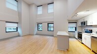 This two-story residence is defined by its dramatic double-height living area, where stacked rows of black-framed windows — positioned at both lower and upper levels — flood the space with natural light and provide views of the surrounding urban streetscape. Light hardwood flooring extends throughout the main level, complementing the neutral warm-white walls and creating a bright, open atmosphere.

The kitchen is efficiently designed with white cabinetry, a full suite of stainless steel appliances including a gas range, microwave, dishwasher, and refrigerator, as well as a central island with a clean white countertop. Track lighting overhead enhances functionality in this area.

A staircase leads to the upper floor, where a hallway with plush carpet connects multiple bedrooms. Each bedroom features carpet flooring, roller-shaded windows with urban views, and ample space. The upper level also includes a bathroom with a glass-enclosed walk-in shower and tile flooring, while a second bathroom on the lower level offers a full soaking tub with tiled surround. The overall aesthetic is clean, contemporary, and well-suited to urban living.