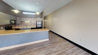 This single-floor unit offers a practical, open layout that flows naturally from a spacious living area into a well-equipped kitchen. The living space is finished with wood-look luxury vinyl flooring throughout and neutral-toned walls, creating a clean and cohesive aesthetic. A bank of three large windows along one wall floods this area with natural light and provides street-level views of the surrounding urban environment, while track lighting overhead ensures ample illumination.

The kitchen is designed for functionality, featuring dark cabinetry with textured finishes, warm-toned countertops, and a full suite of black appliances including a range, microwave, dishwasher, and refrigerator. A peninsula with a blue-accented base extends the counter space and creates a natural separation between the kitchen and living areas. Under-cabinet and overhead fluorescent lighting add practical task illumination.

A separate enclosed room offers a versatile private space, featuring exposed overhead ductwork and a textured ceiling, with the same consistent flooring as the rest of the unit. The bathroom is well-appointed with a sliding glass walk-in shower, a toilet, vanity, and glass shelving, all finished in coordinating neutral tones with chrome hardware accents.