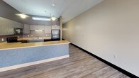 This single-floor unit offers a practical, open layout that flows naturally from a spacious living area into a well-equipped kitchen. The living space is finished with wood-look luxury vinyl flooring throughout and neutral-toned walls, creating a clean and cohesive aesthetic. A bank of three large windows along one wall floods this area with natural light and provides street-level views of the surrounding urban environment, while track lighting overhead ensures ample illumination.

The kitchen is designed for functionality, featuring dark cabinetry with textured finishes, warm-toned countertops, and a full suite of black appliances including a range, microwave, dishwasher, and refrigerator. A peninsula with a blue-accented base extends the counter space and creates a natural separation between the kitchen and living areas. Under-cabinet and overhead fluorescent lighting add practical task illumination.

A separate enclosed room offers a versatile private space, featuring exposed overhead ductwork and a textured ceiling, with the same consistent flooring as the rest of the unit. The bathroom is well-appointed with a sliding glass walk-in shower, a toilet, vanity, and glass shelving, all finished in coordinating neutral tones with chrome hardware accents.