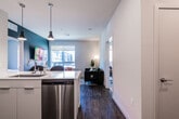 This thoughtfully designed single-floor residence showcases a contemporary aesthetic with a cohesive palette of bold teal accents, crisp white surfaces, and warm wood-look flooring throughout the main living areas. The open-concept kitchen is a standout feature, equipped with sleek white flat-front cabinetry, a generous white quartz peninsula with bar seating, stainless steel appliances including a refrigerator, range, microwave, and dishwasher, and stylish black pendant lighting. The kitchen flows seamlessly into the living area, which benefits from a large window that draws in natural light and offers views of the surrounding urban environment. A teal accent wall adds a bold design element, tying the kitchen and living spaces together. The bedroom is a serene retreat, featuring plush carpeting, a dramatic dark green geometric accent wall, a ceiling fan, and a sizable window with city views that fill the room with daylight. The unit includes two bathrooms: one with a combined tub and shower, white vanity, and wood-look tile flooring, and a second more compact bathroom with a shower. The overall ambiance is modern and urban, with carefully curated design details that create a stylish and comfortable living environment.