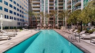 Located at 255 W 5th St in San Pedro, California, this outdoor amenity deck offers a well-appointed communal retreat set against the backdrop of a modern high-rise development. The centerpiece of the space is a pair of resort-style swimming pools — one a long, rectangular lap-style pool lined with rows of chaise lounges and market umbrellas, and a second, more leisurely pool flanked by lounge chairs and mature trees that provide natural shade and privacy.

The surrounding deck is finished in large-format concrete pavers, creating a clean and contemporary aesthetic throughout. A pergola structure on one side of the deck offers a shaded seating area, ideal for relaxing out of direct sunlight. Nearby, a dedicated outdoor kitchen zone features multiple stainless steel BBQ grills alongside a generous concrete patio and a patch of manicured lawn, providing an ideal setting for outdoor gatherings.

Tall palm trees are strategically placed throughout the courtyard, reinforcing the Southern California ambiance, while ornamental shrubs, planters, and green hedgerows add texture and definition to the space. The entire amenity area is enclosed by glass-panel fencing, maintaining open sightlines while ensuring security. The space is bathed in natural sunlight and offers views of the surrounding multi-story residential towers, creating a vibrant, urban-resort atmosphere.